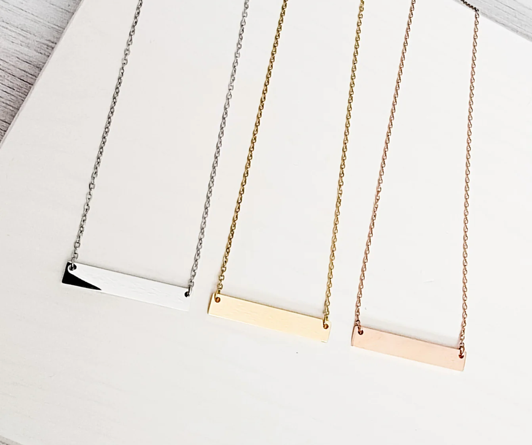 Three necklaces with horizontal bar pendants in silver, gold, and rose gold on a white surface.
